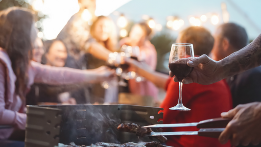Fleisch wird gegrillt mit einem Glas Wein in der Hand und im Hintergrund stoßen Menschen mit ihren Gläsern an