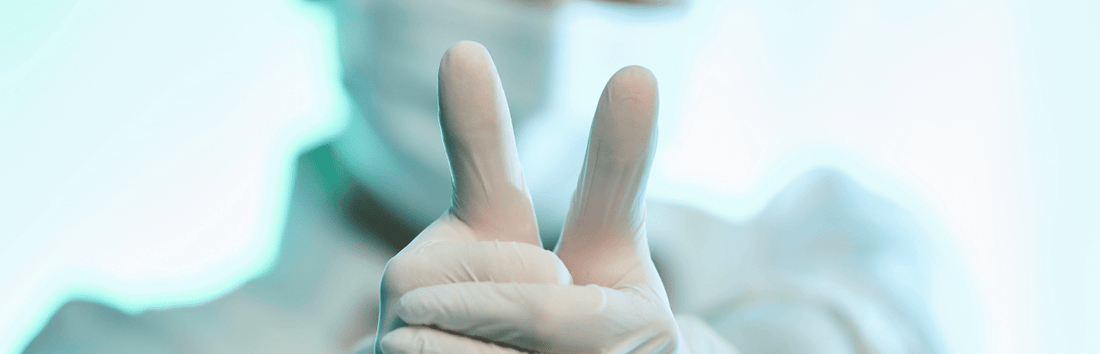 Hand mit medizinischem Handschuh zeigt zwei ausgestreckte Finger, unscharfer klinischer Hintergrund im Bild