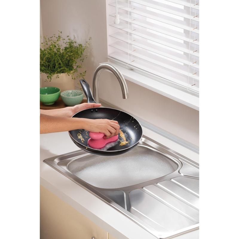 Ένα άτομο καθαρίζει ένα τηγάνι κάτω από τρεχούμενο νερό σε μια σύγχρονη κουζίνα με ένα Scotch-Brite® Delicate Color Comfort μη γρατζουνιστικό σφουγγάρι καθαρισμού (2 τεμάχια) από την 3M Deutschland GmbH - ιδανικό για ευαίσθητες επιφάνειες. Πράσινα μπολ και ένα φυτό βρίσκονται στον πάγκο.