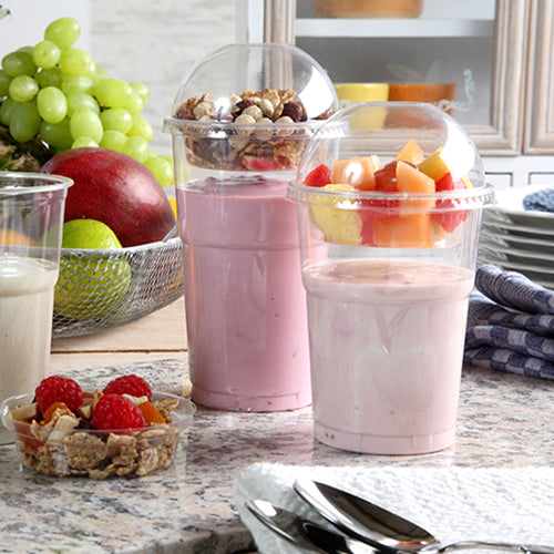 Zwei Joghurtparfaits in Plastikbechern, jeweils verschlossen mit einem Starpak 50 Dom-Deckel „Hurricane“ PET rund glasklar Ø 9,5 cm, stehen auf einer Küchentheke – einer mit Müsli und Nüssen, der andere mit Obst. Im Hintergrund sind frische Beeren und gestapelte Teller zu sehen.