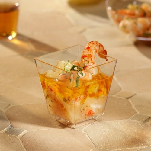 Ένα Starpak 50 Fingerfood-Schale (PS glasklar) της Starpak, γεμάτο με γαρίδες και κοκτέιλ θαλασσινών και γαρνιρισμένο με βότανα, βρίσκεται σε μπεζ, υφή επιφάνεια. Άλλα ποτήρια είναι θολά στο φόντο – τέλεια για στυλάτες εκδηλώσεις.