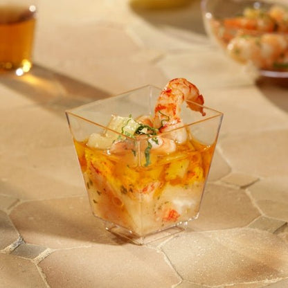 Ένα Starpak 50 Fingerfood-Schale (PS glasklar) της Starpak, γεμάτο με γαρίδες και κοκτέιλ θαλασσινών και γαρνιρισμένο με βότανα, βρίσκεται σε μπεζ, υφή επιφάνεια. Άλλα ποτήρια είναι θολά στο φόντο – τέλεια για στυλάτες εκδηλώσεις.