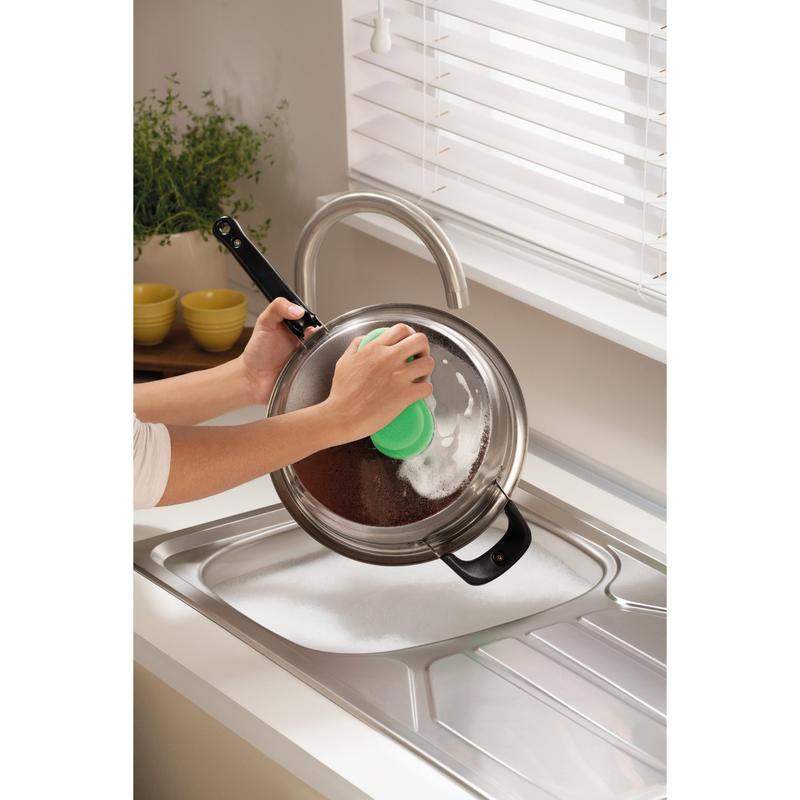 Κάποιος καθαρίζει ένα μεταλλικό καπάκι κατσαρόλας πάνω από έναν σαπουνισμένο νεροχύτη με το Scotch-Brite® Classic Reinigungsschwamm της 3M Deutschland GmbH, κοντά σε ένα παράθυρο με λευκές περσίδες και έναν πάγκο με ένα φυτό και στοιβαγμένα κίτρινα μπολ.