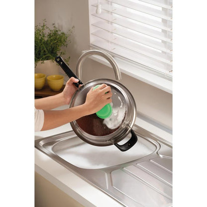 Κάποιος καθαρίζει ένα μεταλλικό καπάκι κατσαρόλας πάνω από έναν σαπουνισμένο νεροχύτη με το Scotch-Brite® Classic Reinigungsschwamm της 3M Deutschland GmbH, κοντά σε ένα παράθυρο με λευκές περσίδες και έναν πάγκο με ένα φυτό και στοιβαγμένα κίτρινα μπολ.