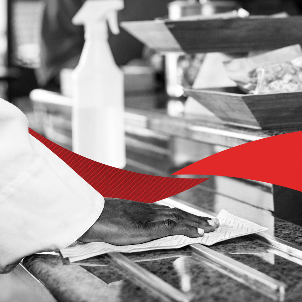 Eine Hand reinigt eine Granitarbeitsplatte mit WypAll® L20 Papierwischtuch (2-lagig, 6 x 550 Blatt) von Kimberly-Clark GmbH; dahinter sind eine Sprühflasche und Lebensmittelschalen zu sehen, mit einem roten Grafikelement quer über dem Bild.