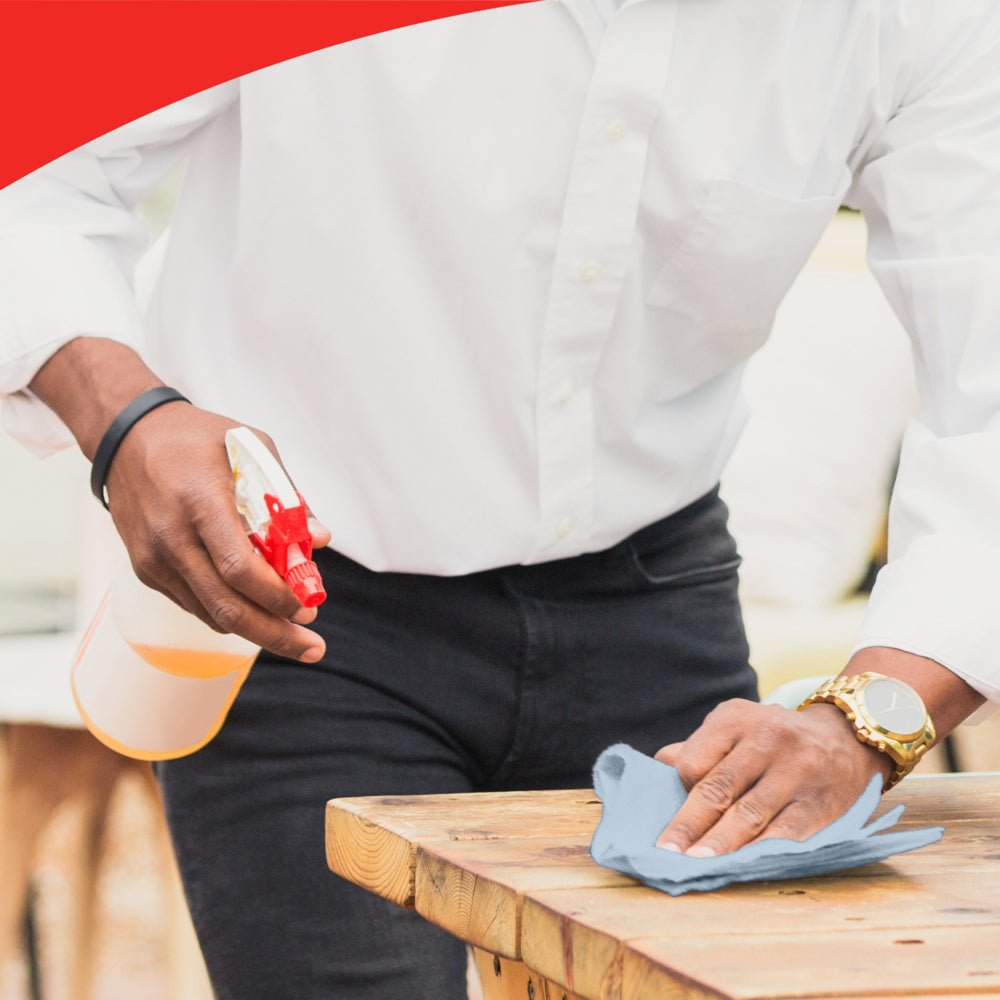 Ένα άτομο με λευκό πουκάμισο και ρολόι καθαρίζει ένα ξύλινο τραπέζι με ένα μπλε πανί, πιθανώς WypAll® L20 General Clean - BRAG™ Box της Kimberly-Clark GmbH. Η εικόνα έχει πάνω μια κόκκινη καμπύλη άκρη που τονίζει την προσοχή στη λεπτομέρεια.