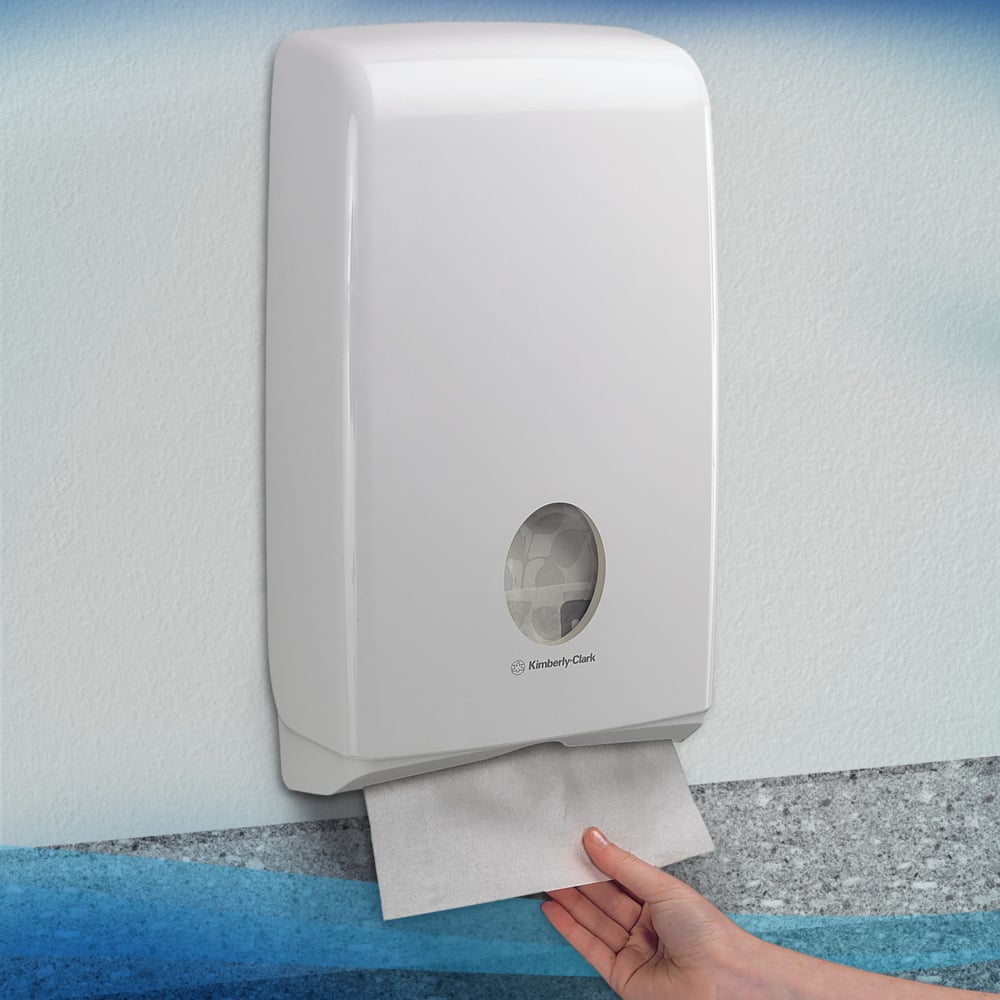 Ένα χέρι τραβάει μια Scott® SLIMFOLD χαρτοπετσέτα από έναν λευκό τοίχο διανομέα της Kimberly-Clark GmbH με παράθυρο προβολής, που δείχνει διπλωμένες χαρτοπετσέτες στο εσωτερικό. Ο υγιεινός διανομέας είναι τοποθετημένος σε έναν φωτεινό τοίχο. Χωράει 16 κλιπ x 110 φύλλα ανά κουτί.