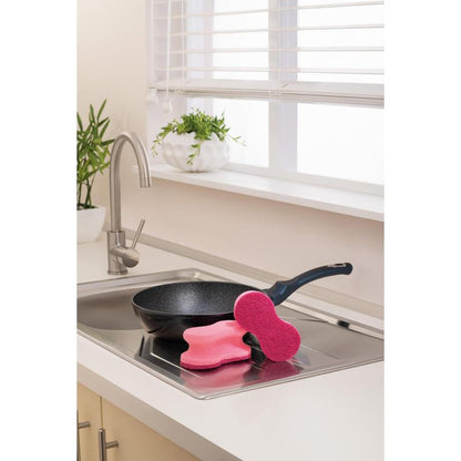 Ένα μαύρο τηγάνι βρίσκεται σε έναν νεροχύτη κουζίνας, στο χείλος του οποίου βρίσκονται δύο ροζ Scotch-Brite® Delicate Color Comfort μη γρατζουνιστικό σφουγγάρι καθαρισμού (2 τεμάχια, 3M Deutschland GmbH). Ο λευκός πάγκος και τα φυτά σε γλάστρες κοντά ολοκληρώνουν τη σκηνή.