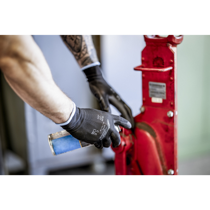 Ένα άτομο με νάιλον γάντια NITRAS, μαύρα, επικαλυμμένα με PU από την AS Arbeitsschutz GmbH καθαρίζει ή λιπαίνει με ένα μπλε δοχείο ψεκασμού ένα κόκκινο μεταλλικό εξάρτημα μηχανής. Φαίνονται μόνο τα χέρια και οι αγκώνες.