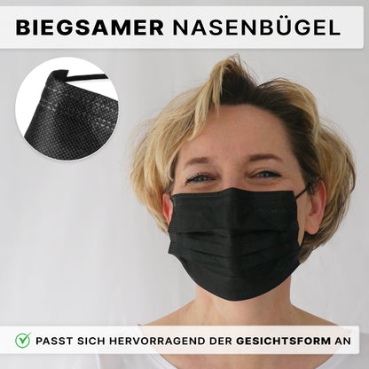 Eine Person mit kurzen blonden Haaren trägt einen ARNOMED MUNDSCHUTZ TYP IIR OP-Mundschutz mit Gummibändern der ARNOWA GmbH. Die schwarze Einwegmaske mit flexiblem Nasenbügel ist in einer Nahaufnahme zu sehen, mit deutschem Text, der auf die verstellbare Passform hinweist.