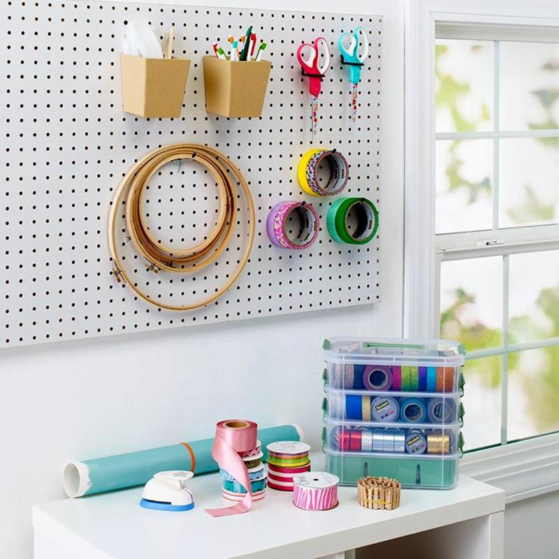 Ένας χώρος χειροτεχνίας με μια πλάκα καρφιών, στην οποία κρέμεται το κόκκινο ψαλίδι ακρίβειας Scotch™ από την 3M Deutschland GmbH, στεφάνες κεντήματος, ρολά ταινίας και στυλό. Κάτω είναι διατεταγμένα υλικά χειροτεχνίας και κορδέλες σε ένα λευκό τραπέζι κοντά σε ένα παράθυρο.