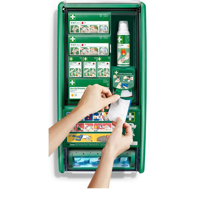 Eine Person hält einen Brandgel-Verband vor der Cederroth Erste Hilfe & Verbrennungsstation (1 Packung) von Orkla Wound Care AB, einer grünen Erste-Hilfe-Station mit Verbrennungsmaterial, Verbänden, Flaschen und Anweisungen.