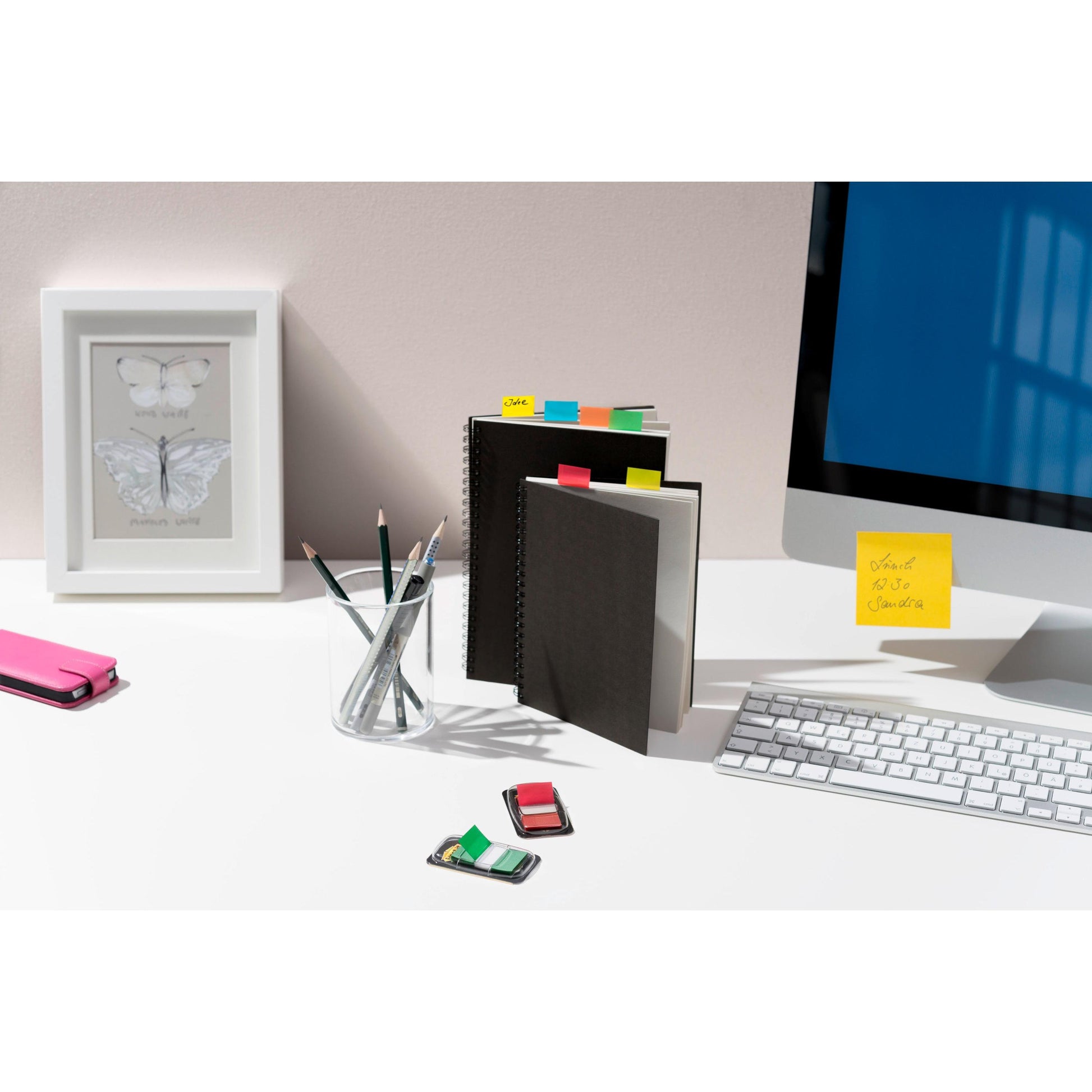 Ein aufgeräumter Schreibtisch mit Monitor, Tastatur, Handytasche, Bleistiften in einem Glas, Radiergummis, gerahmtem Schmetterlingsdruck und geschlossenen Notizbüchern, die mit Post-it® Index Haftstreifen der 3M Deutschland GmbH (25,4x43,2mm, 50 Streifen/Spender) beschriftet sind.