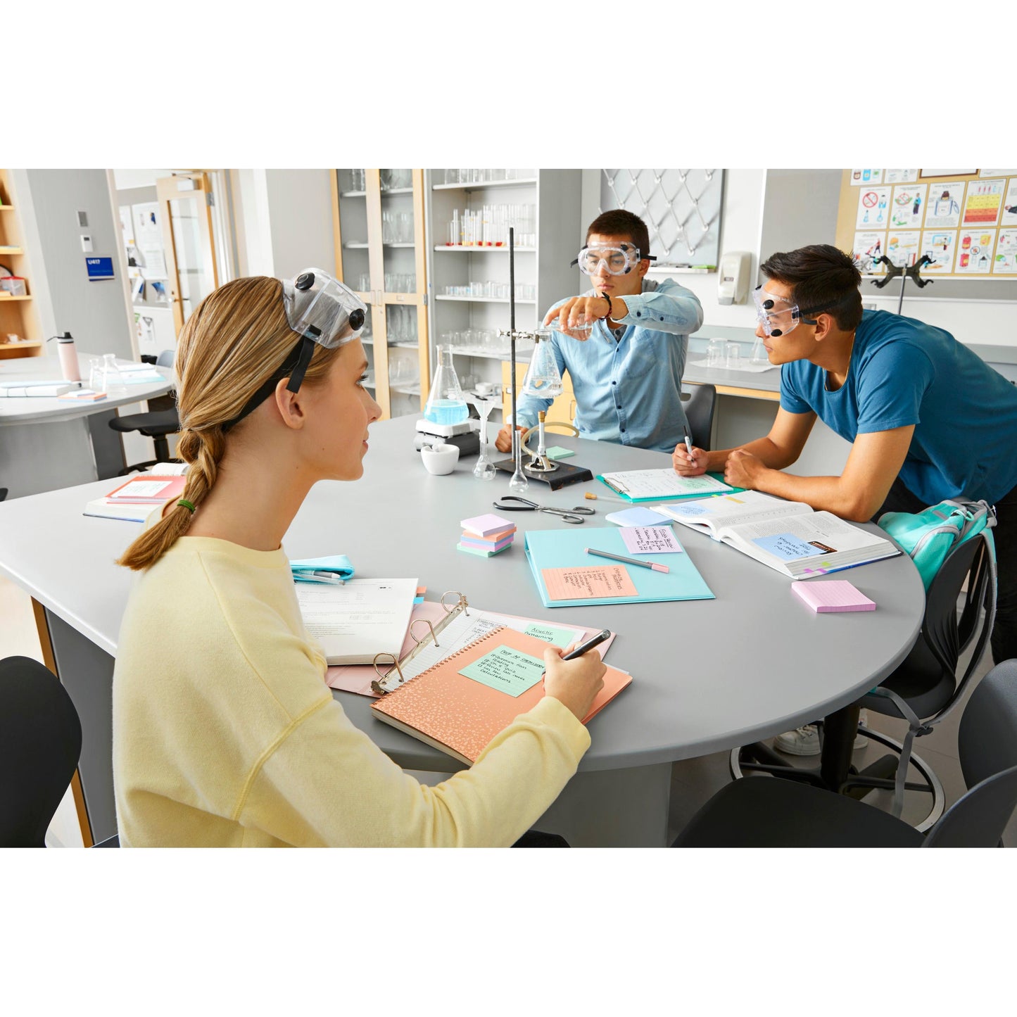 Drei Studenten mit Schutzbrillen sitzen an einem runden Labortisch mit Notizbüchern, Lehrbüchern und Post-it® Super Sticky Notes (76 mm x 76 mm, 90 Blatt/Block, 100% PEFC) von 3M Deutschland GmbH und führen ein Experiment mit einem Stativ und einem Becherglas durch.