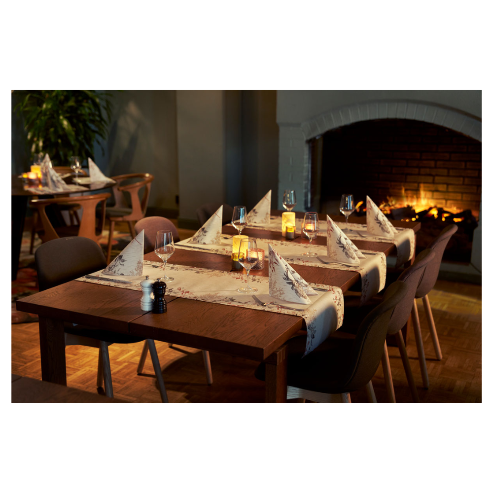 Ένας άνετος χώρος τραπεζαρίας διαθέτει δύο ξύλινα τραπέζια με ποτήρια κρασιού και πετσέτες Duni Bio Dunisoft® δίπλα σε τρεμοπαίζουσες κεριά. Σκούρες καρέκλες περιβάλλουν τα τραπέζια, αναβαθμισμένες από ένα αναμμένο τζάκι. Απαλές θέσεις και ένα φυτό συμβάλλουν στο σχέδιο εμπνευσμένο από τη φύση.