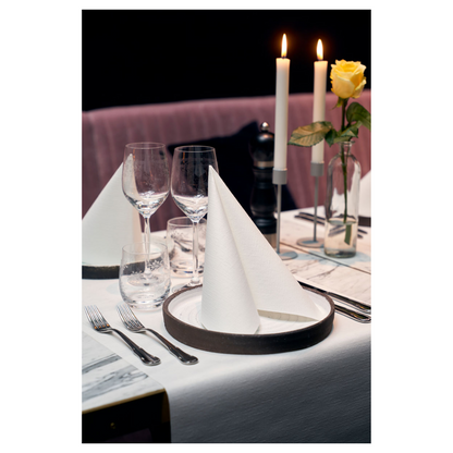 Ένα τραπέζι δείπνου είναι κομψά στρωμένο με δύο χαρτοπετσέτες Duni Dunilin® 48 x 48 cm Λευκές, που είναι διπλωμένες σε τρίγωνα πάνω στα πιάτα. Δύο μεγάλες αναμμένες κεριά και ένα κίτρινο τριαντάφυλλο σε γυάλινο βάζο κοσμούν το φόντο. Ποτήρια κρασιού, ένα ποτήρι νερού και μαχαιροπήρουνα συμπληρώνουν αυτό το απαιτητικό περιβάλλον με υφή που μοιάζει με ύφασμα.