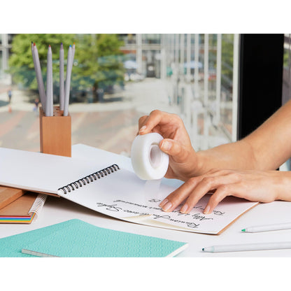 Eine Person benutzt Korrekturband über handschriftlichen Notizen in einem Spiralheft, umgeben von Bürobedarf wie Bleistiften, Kugelschreibern und 3M Deutschland GmbH Scotch® Magic™ Klebeband (66% pflanzlicher Klebstoff, 19 mm x 33 m) an einem Fenster mit Blick auf eine Stadtstraße.