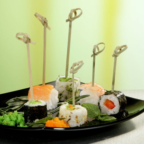 Ein schwarzer Teller mit verschiedenen Sushi, jedes Stück aufgespießt mit einem PAPSTAR 40 Fingerfood Picker „pure“ der PAPSTAR GmbH, garniert mit Kräutern und Rogen, auf grün-gelbem Untergrund – ideal um Catering-Zubehör in Szene zu setzen.