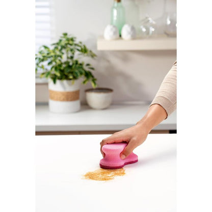 Ένα χέρι χρησιμοποιεί ένα ροζ Scotch-Brite® Delicate Color Comfort μη γρατζουνιστικό σφουγγάρι καθαρισμού (2 τεμάχια, 3M Deutschland GmbH) για να καθαρίσει καφέ σκόνη από έναν λευκό πάγκο - τέλειο για ευαίσθητες επιφάνειες. Φυτό και μπολ στο φόντο.