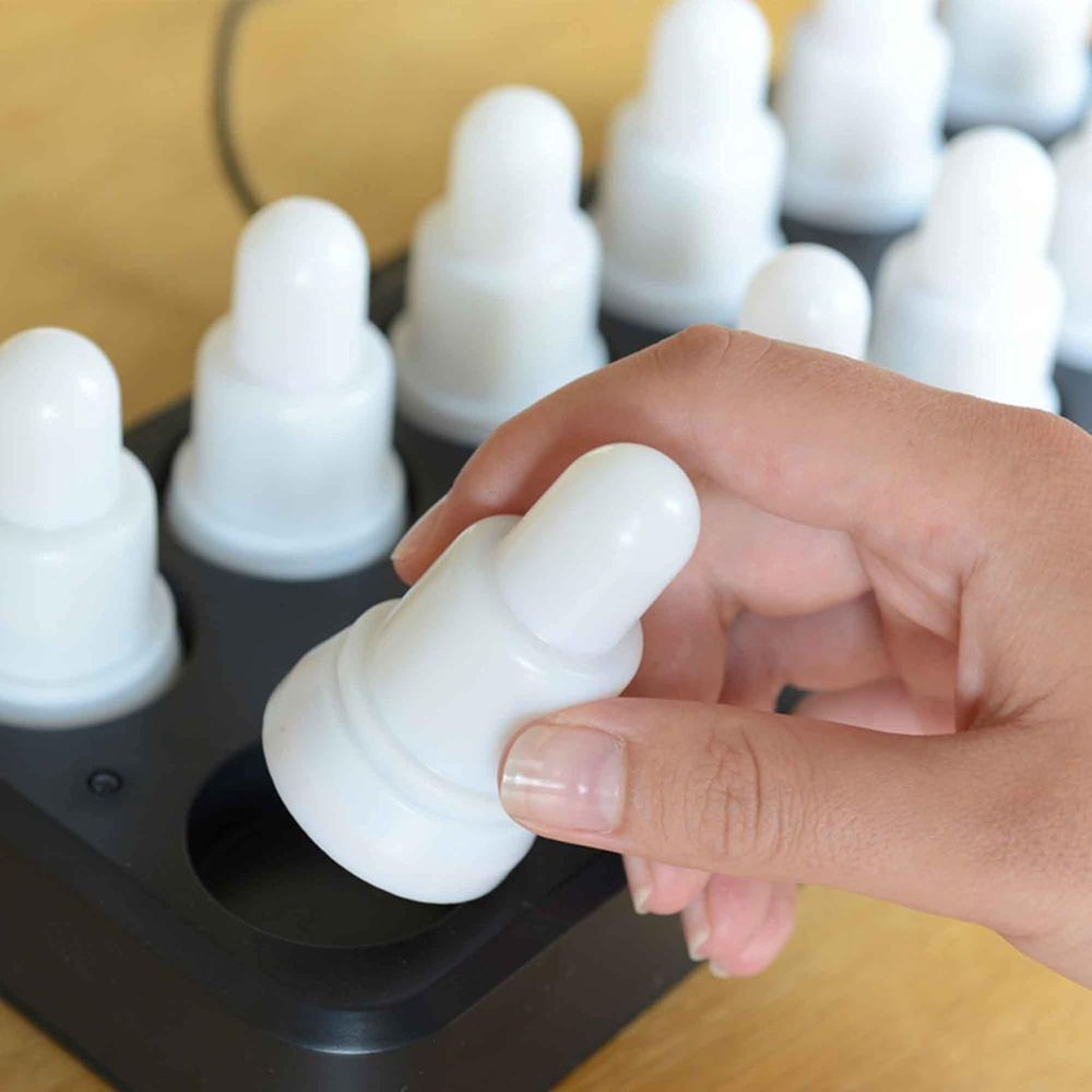 Ένα χέρι τοποθετεί ή αφαιρεί ένα λευκό, στρογγυλό LED κερί από το μαύρο δίσκο του συνόλου LED της Duni GmbH, Θερμό Λευκό & Πολύχρωμο - 12 τεμάχια φώτα αλλαγής χρώματος; κάθε πανομοιότυπο, επαναφορτιζόμενο κερί κάθεται σε ξύλινη βάση.