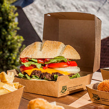 Ein Cheeseburger mit Salat, Tomate, Zwiebel und Cheddar-Käse liegt im Freien in einer PAPSTAR GmbH Burgerbox aus Pappe (9x18,5x18,5 cm, "100% Fair", 25 Stück). Pommes frites und andere Beilagen werden separat daneben serviert.