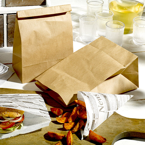PAPSTAR 250 Blockbodenbeutel aus braunem Kraftpapier von PAPSTAR GmbH stehen mit Zeitungspapier-verpacktem Essen auf Holz, dazu Gläser, ein Krug mit hellem Getränk sowie Pommes und ein Burger im Hintergrund.