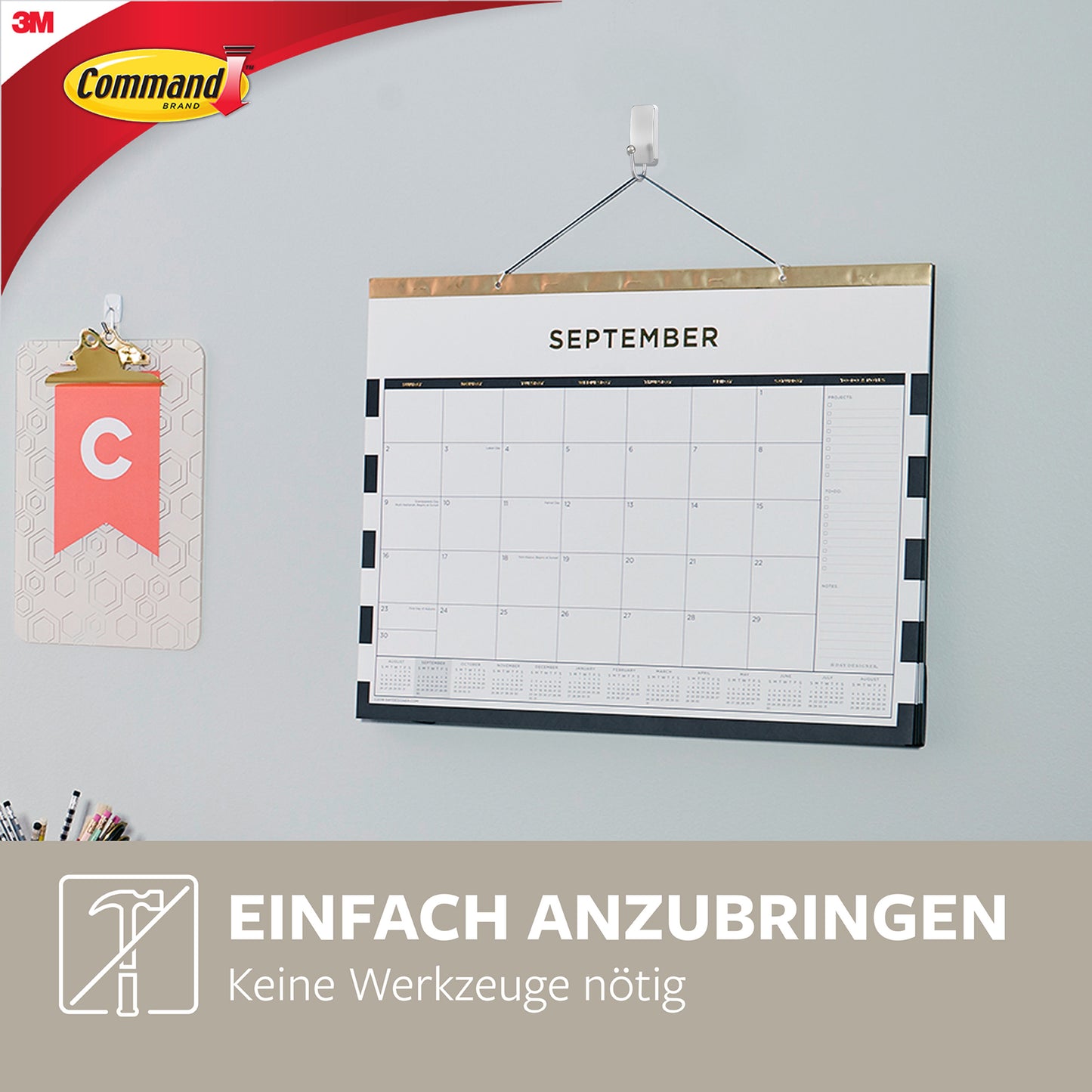 Ein kleiner Command™ Metallhaken in gebürstetem Nickel von 3M Deutschland GmbH hält einen Wandkalender an einer hellgrauen Wand. Daneben hängt ein kleines Klemmbrett mit dem Buchstaben "C". Einfach anzubringen, keine Werkzeuge nötig.