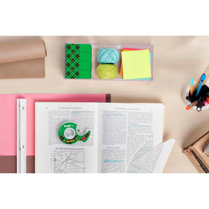 Ένας χώρος εργασίας με ένα ανοιχτό βιβλίο, χιλιομετρικό χαρτί, χρωματιστές μπάλες νήματος, αυτοκόλλητα σημειώματα, στυλό σε ποτήρι, γόμες, μια καφέ ρολό χαρτί συσκευασίας και ένα ρολό 3M Deutschland GmbH Scotch® Magic™ Αόρατης Ταινίας σε ανοιχτό ξύλινο τραπέζι.