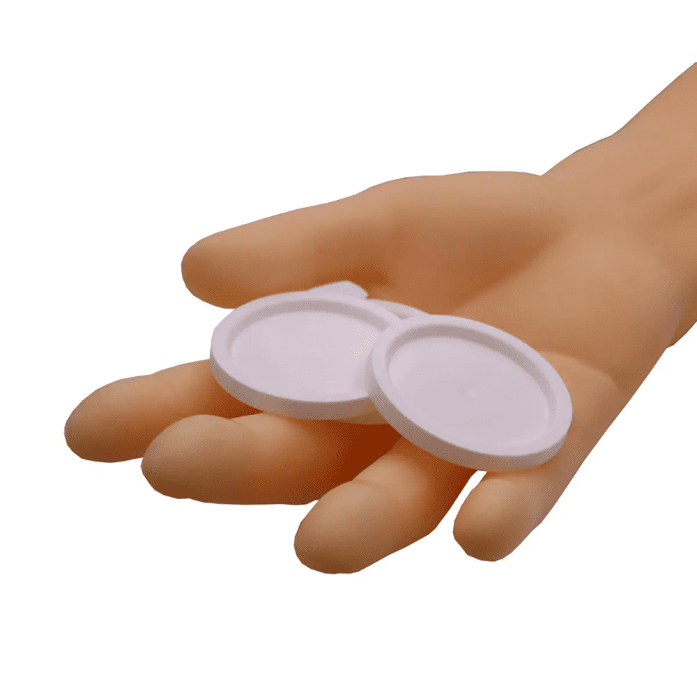 Ένα χέρι κρατάει δύο στρογγυλά καπάκια AMPri για ιατρικά ποτήρια, λευκά αντικείμενα μπροστά από ένα απλό λευκό φόντο.