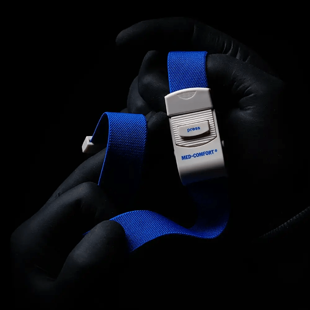 Ένα άτομο με μαύρα γάντια κρατάει μια μπλε AMPri MED-COMFORT φλεβική δέστρα, ένα απαραίτητο εργαλείο για ιατρικές εφαρμογές. Σε σκοτεινό φόντο διακρίνεται μια λευκή πλαστική αγκράφα με κουμπί πίεσης.