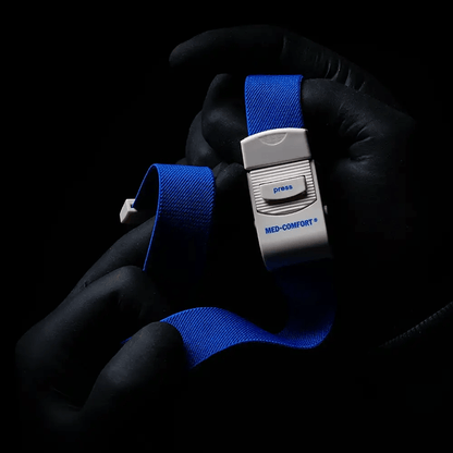 Ένα άτομο με μαύρα γάντια κρατάει μια μπλε AMPri MED-COMFORT φλεβική δέστρα, ένα απαραίτητο εργαλείο για ιατρικές εφαρμογές. Σε σκοτεινό φόντο διακρίνεται μια λευκή πλαστική αγκράφα με κουμπί πίεσης.