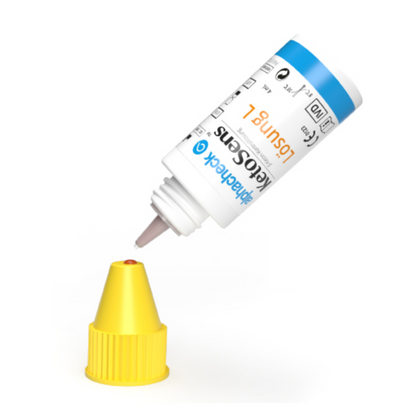 Ένα λευκό μπουκάλι με μπλε-κίτρινη ετικέτα είναι αναποδογυρισμένο, με το ακροφύσιο στάλαξης να βρίσκεται πάνω από ένα κίτρινο καπάκι. Η ετικέτα περιέχει κείμενο και σύμβολα που υποδεικνύουν ότι πιθανώς πρόκειται για προϊόν της λύσης ελέγχου β-κετόνης Alphacheck KetoSens L+M | Συσκευασία (2 φιαλίδια) από την Berger Med GmbH για χρήση με συσκευές μέτρησης κετόνης.