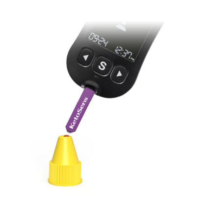 Μια ψηφιακή συσκευή μέτρησης με ένδειξη «09:24» και «12:37 μ.μ.» χρησιμοποιείται για να δοκιμάσει μια μοβ λωρίδα με την επιγραφή «Alphacheck KetoSens β-κετόνη λύση ελέγχου L+M | Συσκευασία (2 φιαλίδια)». Η λωρίδα Alphacheck δείχνει σε ένα μικρό κίτρινο καπάκι. Η συσκευή διαθέτει ένα μεγάλο κουμπί «S» κάτω από την ένδειξη.