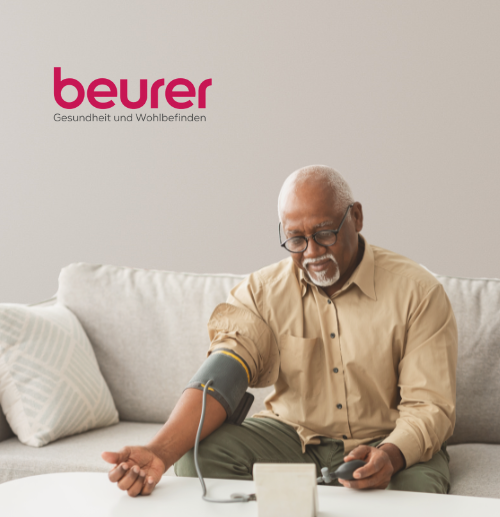 Ein älterer Mann sitzt auf einem weißen Sofa und verwendet ein Blutdruckmessgerät für den Oberarm. Er trägt eine Brille und ein beiges Hemd. Der Hintergrund ist minimalistisch mit hellen Wänden und einem Kissen. In der oberen linken Ecke des Bildes steht der Text „beurer Gesundheit und Wohlbefinden“.
