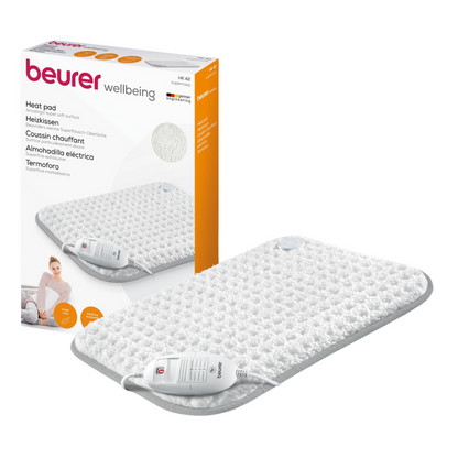 Δίπλα στο προϊόν απεικονίζεται ένα Beurer HK 42 θερμαινόμενο μαξιλάρι Super-Cosy της Beurer GmbH με τη μάρκα «wellbeing» στη συσκευασία. Το λευκό μαξιλάρι με γκρι περίγραμμα είναι εξοπλισμένο με τηλεχειριστήριο. Η συσκευασία δείχνει επίσης μια εικόνα του θερμαινόμενου μαξιλαριού σε χρήση και τονίζει τη λειτουργία γρήγορης θέρμανσης.