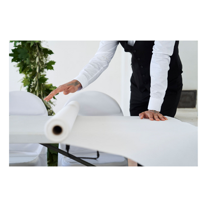 Ένα άτομο με βραδινή ενδυμασία, με λευκό πουκάμισο και μαύρο γιλέκο, ξετυλίγει ένα ρολό τραπεζομάντιλου Dunicel (1,18 μ. x 40 μ.) της Duni GmbH πάνω σε ένα τραπέζι. Στο απλό φόντο φαίνεται λίγο πράσινο.