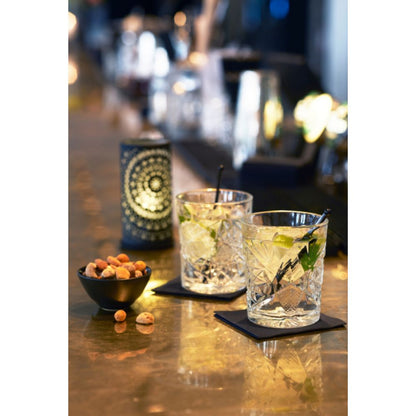 Δύο ποτήρια με πάγο, φέτες λάιμ και καλαμάκια βρίσκονται σε τετράγωνες σουβέρ πάνω σε μια μπάρα. Δίπλα τους υπάρχει ένα μικρό μαύρο μπολ με ανάμεικτους ξηρούς καρπούς. Η μεταλλική βάση κεριών Billy της Duni GmbH, σε απλό μέταλλο 140 x 75 mm με μοντέρνο σχεδιασμό, φωτίζει απαλά στο παρασκήνιο, δημιουργώντας μια ζεστή ατμόσφαιρα.