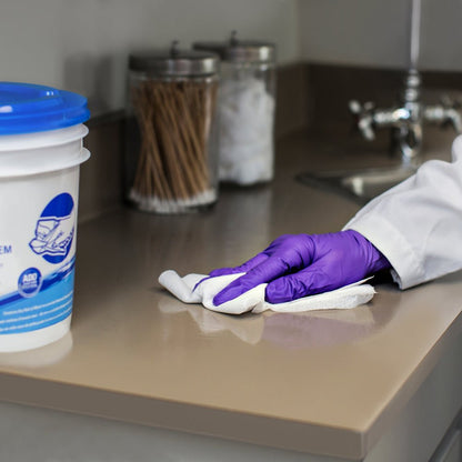 Ένα άτομο με μωβ γάντια σκουπίζει με υφάσματα καθαρισμού WypAll® Wettask™ της Kimberly-Clark GmbH (6 ρόλοι x 90 φύλλα) μια μπεζ επιφάνεια εργασίας κοντά στο νεροχύτη, ενώ στο φόντο φαίνονται βάζα με βαμβακοφόρους στυλεούς και μπάλες.