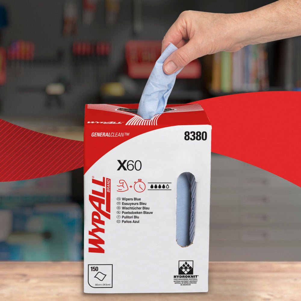 Ένα χέρι τραβάει ένα μπλε φύλλο WypAll® X60 General Clean από ένα λευκό-κόκκινο κουτί διανομής της Kimberly-Clark GmbH με την επιγραφή "8380", "150 φύλλα" και "Κεντρική διάθεση". Στο φόντο φαίνεται ένα θολό εργαστήριο με ράφια.