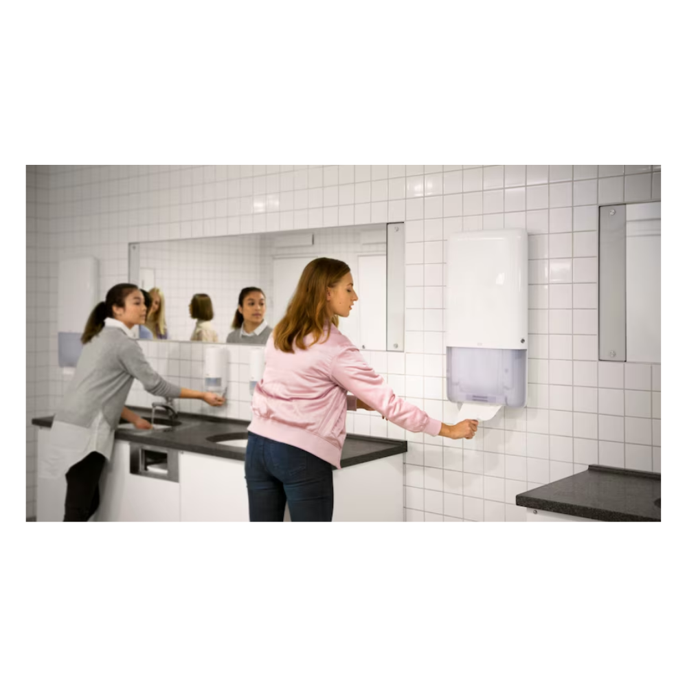 Zwei Frauen stehen in einer öffentlichen Toilette. Eine Frau in einer rosa Jacke nimmt ein Papierhandtuch aus einem Tork PeakServe® 552500 Spender für Endlos Handtücher Elevation H5 | Karton (1 Packungen) von TORK, während die andere Frau in einem grauen Pullover ihre Hände an einem Waschbecken wäscht. Die Toilette, die für hochfrequentierte Waschräume geeignet ist, hat weiß geflieste Wände und große Spiegel.