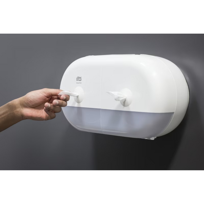 Eine Person zieht ein Papierhandtuch aus einem eleganten weißen Tork SmartOne® 682000 Mini Doppelrollenspender für Toilettenpapier, der über zwei Fächer verfügt und an einer glatten, dunkelgrauen Wand angebracht ist.