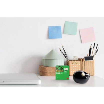Ένα γραφείο με έναν κλειστό φορητό υπολογιστή, 3M Scotch™ Magic Tape, ένας μαύρος επιτραπέζιος διανομέας Scotch™ Karim Rashid της 3M Deutschland GmbH, κούπα με στυλό και μολύβια, στοιβαγμένα στρογγυλά κουτιά και τρία παστέλ αυτοκόλλητα σημειώματα σε έναν λευκό τοίχο.