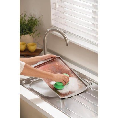 Κάποιος τρίβει ένα μεγάλο, βρώμικο ταψί με το Scotch-Brite® Classic Reinigungsschwamm της 3M Deutschland GmbH κάτω από μια βρύση σε μια φωτεινή κουζίνα με λευκές περσίδες, ένα φυτό και στοιβαγμένα κίτρινα μπολ στο παρασκήνιο.