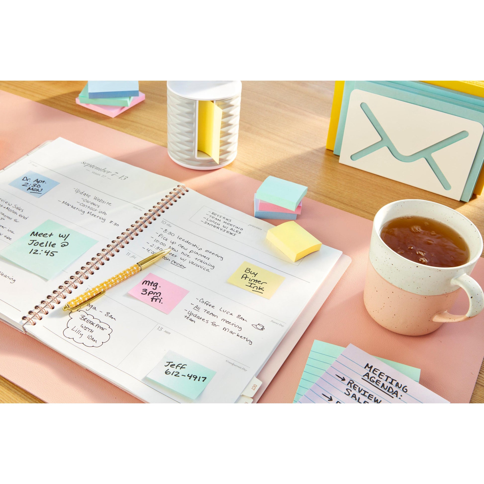 Ein aufgeschlagener Planer mit handschriftlichen Notizen und einem Stift liegt auf einem Schreibtisch mit Post-it® Recycling Notes (76 mm x 76 mm, 100 Blatt/Block, 100% PEFC) von 3M Deutschland GmbH, einer Tasse Tee, einem Karteikarten-Organizer, einer kleinen Lampe und "Meeting Agenda"-Karten.