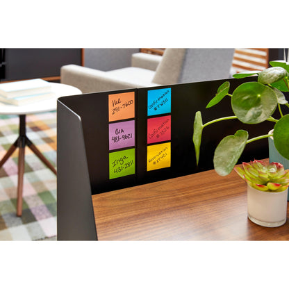 Ein Post-it® Super Sticky Würfel, farbig (440 Blatt, 76 x 76 mm, 100% PEFC) der 3M Deutschland GmbH hält Namen und Telefonnummern neben einem Holzschreibtisch mit Pflanzen; im Hintergrund stehen Bücher und moderne Sitzmöbel.