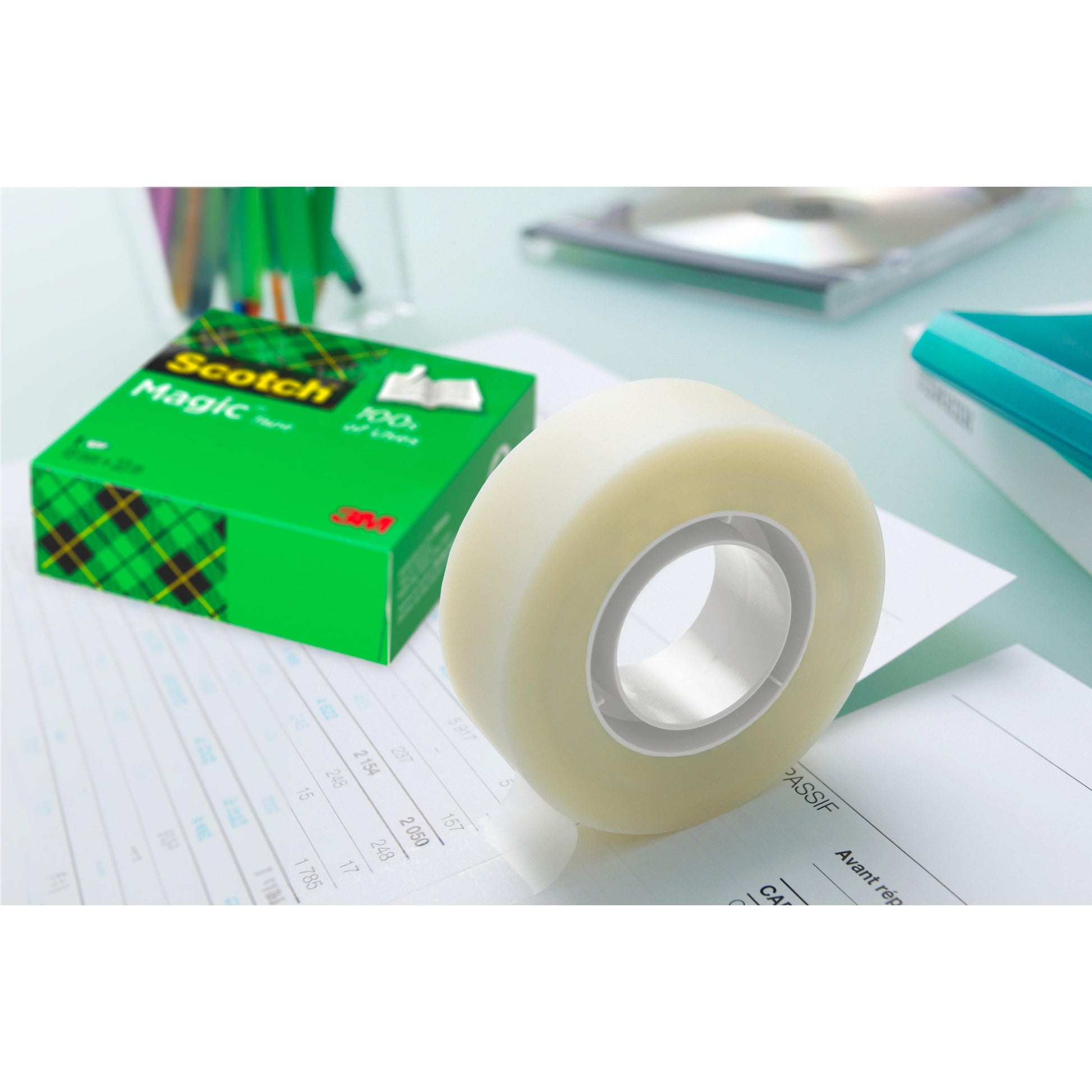 Eine Packung Scotch® Magic™ Unsichtbares Klebeband (8 Rollen, 19 mm x 33 m) von 3M Deutschland GmbH liegt auf einem Dokument, die grüne Schachtel daneben. Stifte, ein Notizbuch und andere Büromaterialien erscheinen unscharf im Hintergrund.